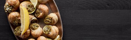 top view of delicious cooked escargots with lemon on black wooden table, panoramic shotの写真素材