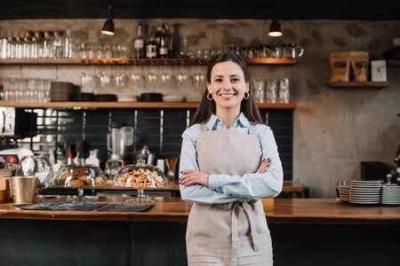 smiling barista in apron standing with crossed arms near bar counterの写真素材
