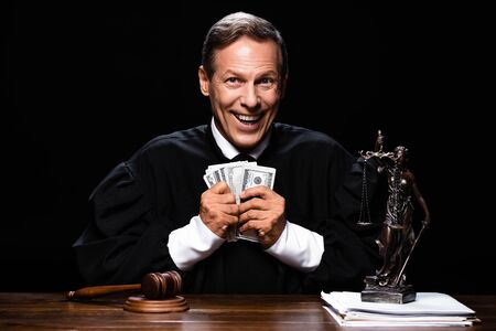 smiling judge in judicial robe sitting at table and holding dollar banknotes isolated on blackの写真素材