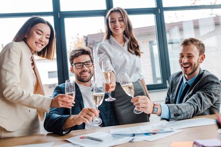cheerful multicultural businesswomen and businessmen holding champagne glasses in officeの写真素材