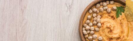 top view of delicious hummus with chickpeas and nacho in bowl on beige wooden table, panoramic shot