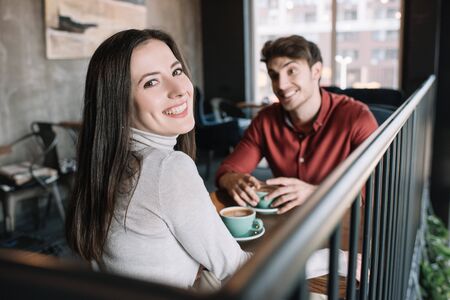 selective focus of smiling girl looking at camera while drinking coffee with man on balcony in coffee shopの写真素材