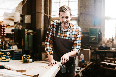 selective focus of happy bearded carpenter carving wood in workshopの写真素材
