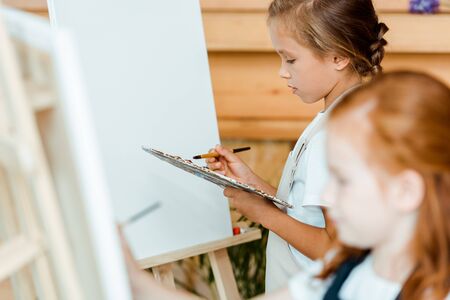 selective focus of cute kid holding palette near easel and childの写真素材