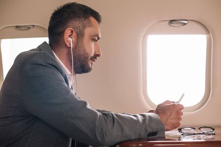 side view of businessman in earphones using smartphone in private jet