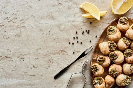 top view of delicious cooked escargots with lemon slices, black peppercorn and tweezers on stone...