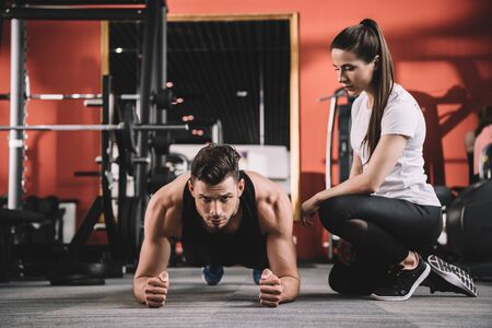 attentive trainer supervising handsome sportsman doing plank exerciseの写真素材