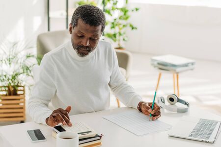 serious african american man sitting at table near utility bill and calculating expensesの写真素材