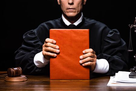 cropped view of judge in judicial robe sitting at table and holding orange book isolated on blackの写真素材