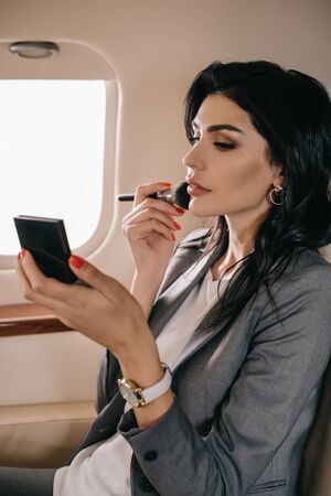 attractive businesswoman holding cosmetic brush near face in private jet