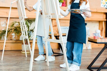 cropped view of kids in aprons standing near easelsの写真素材