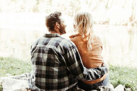 back view of young man embracing girlfriend while sitting near lake in parkの写真素材