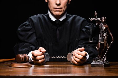 cropped view of judge in judicial robe sitting at table with handcuffs isolated on black