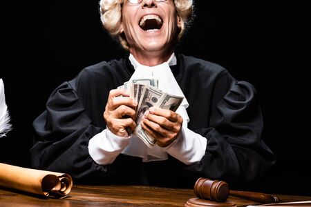 cropped view of smiling judge in judicial robe and wig sitting at table and holding money isolated on blackの写真素材