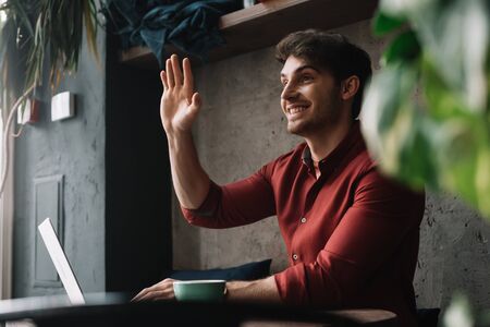 smiling young freelancer working on laptop and waving hand in coffee shopの写真素材