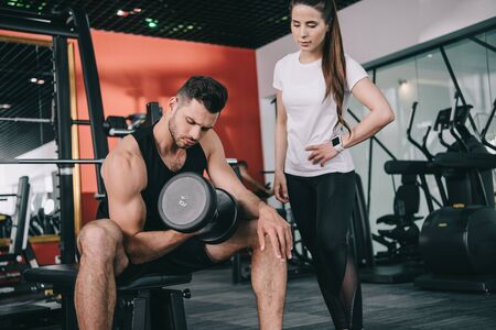 attentive trainer standing near handsome sportsman working out with dumbbellの写真素材