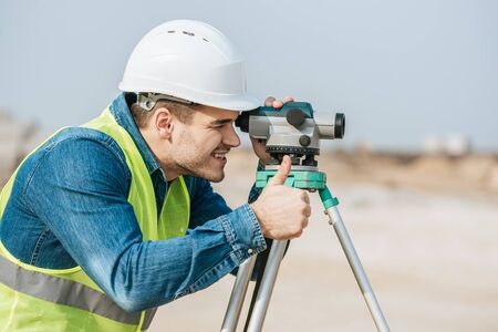 Smiling surveyor looking through digital level and showing thumb up gestureの写真素材