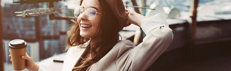 smiling fashionable girl in beige suit and beret posing on roof with coffee to goの写真素材