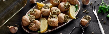 delicious cooked escargots with lemon slices on black wooden table with spices and white wine, panoramic shotの写真素材