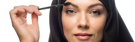 young Muslim woman in hijab applying mascara on eyelashes isolated on white, panoramic shot