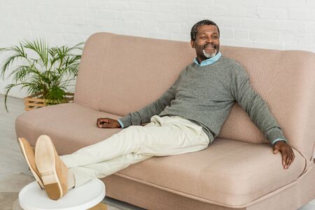 cheerful african american sitting on sofa, putting legs on table and looking awayの写真素材