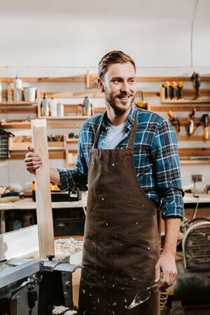 happy carpenter in apron standing and holding wooden plank in workshopの写真素材