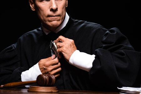 cropped view of scared judge in judicial robe sitting at table and holding dollar banknotes isolated...