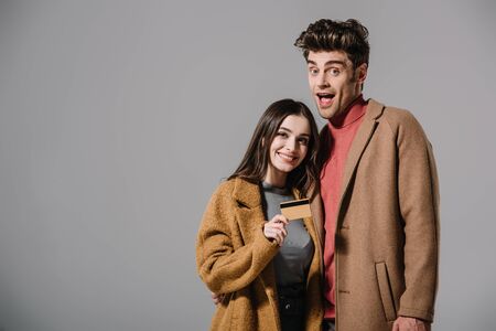 excited couple in beige coats holding credit card isolated on greyの写真素材