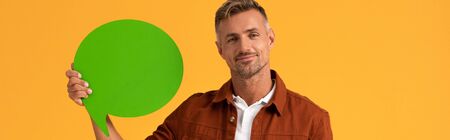 panoramic shot of happy man holding green speech bubble isolated on orange
