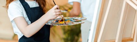 panoramic shot of kid holding palette and paintbrush near easelの写真素材