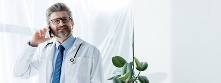 panoramic shot of smiling doctor in white coat talking on smartphone in hospitalの写真素材