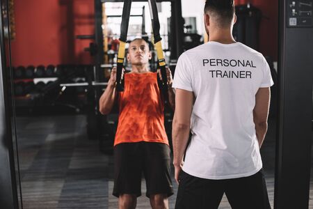 back view of personal trainer standing near young african american athlete pulling up on suspension trainerの写真素材