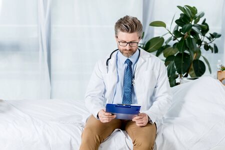 handsome doctor in white coat sitting on bed and looking at clipboard in hospital