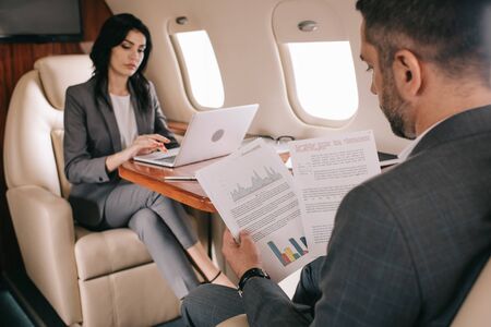 selective focus of bearded businessman holding charts and graphs near attractive businesswoman using laptop in planeの写真素材