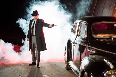 stylish gangster in hat holding gun near retro car and smokeの写真素材