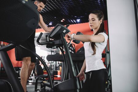 attentive trainer looking at fitness tracker while standing sportsman running on treadmillの写真素材