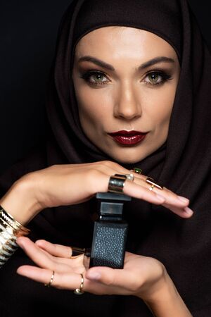 beautiful Muslim woman in hijab with makeup in golden jewelry holding perfume isolated on black
