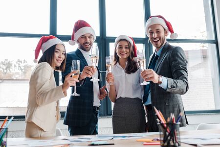 selective focus of happy multicultural businesswomen and businessmen in santa hats holding champagne glasses in officeの写真素材
