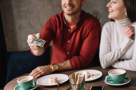 cropped view of young couple eating cheesecake and drinking coffee while man holding money in coffee...