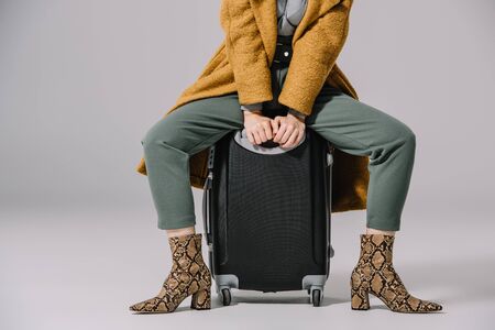 cropped view of stylish woman in beige coat sitting on travel bag on greyの写真素材