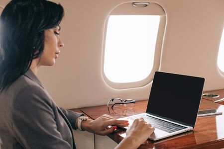 selective focus of attractive businesswoman using laptop with blank screen in private jet