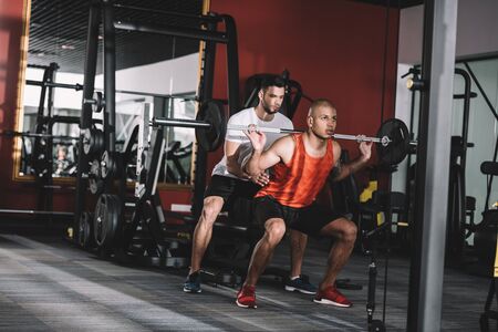 attentive trainer supporting young african american sportsman lifting barbell
