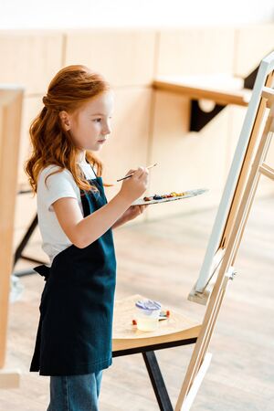 cute redhead kid with paintbrush and palette near easelの写真素材