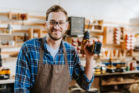 happy carpenter in goggles and apron holding hammer drillの写真素材