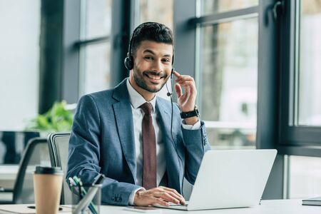 cheerful call center operator looking at camera while sitting at workplace near laptop