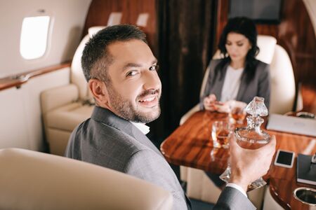 selective focus of happy businessman holding jar with alcohol drink near businesswoman in private...