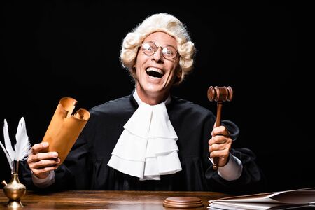 smiling judge in judicial robe and wig sitting at table and holding document and gavel isolated on blackの写真素材