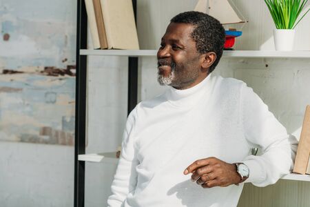 stylish, senior african american man standing near rack, smiling and looking away
