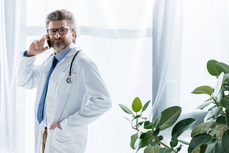 handsome doctor in white coat with hand in pocket talking on smartphone in hospitalの写真素材