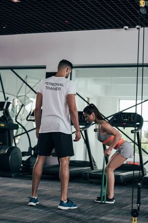 back view of personal trainer looking at athletic sportswoman exercising with resistance bandsの写真素材